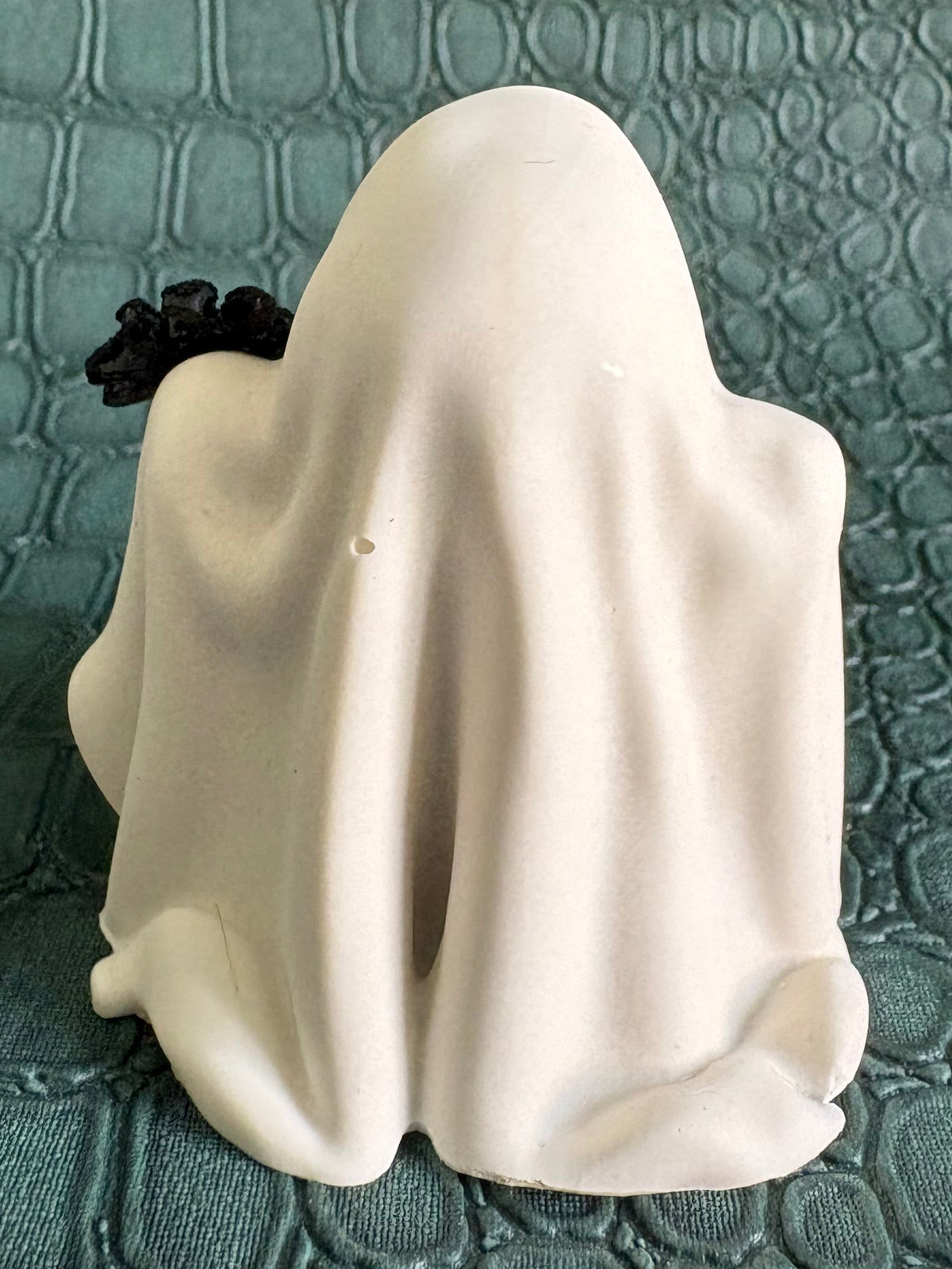 A small white ghost figurine with a black rose accent on its left side, set against a textured teal-green background.