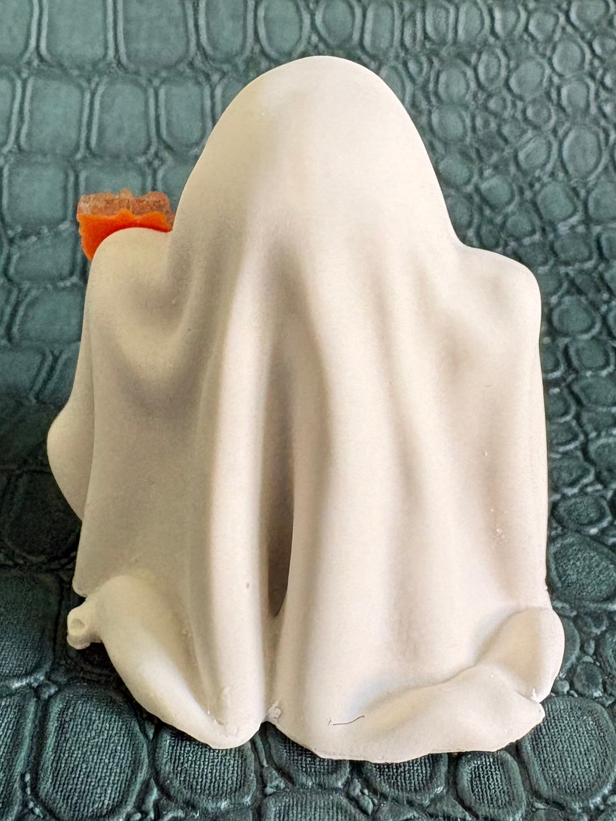 A small white ghost figurine with a bright orange-red accent peeking out from behind it, placed on a dark textured crocodile-patterned surface.