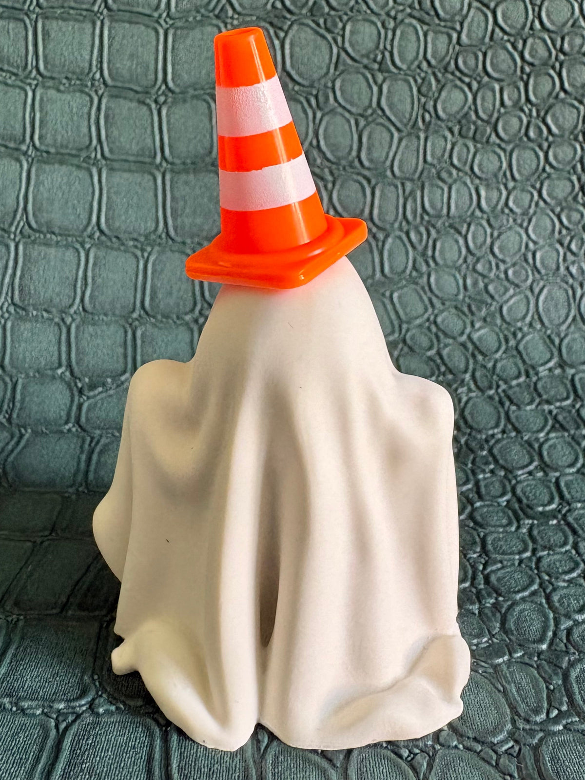 A small Halloween-style ghost figurine draped in an off-white sheet, wearing an orange traffic cone hat, standing on a textured dark green surface.