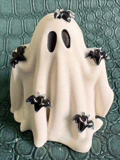 A small white ghost-shaped figurine or candle with black oval eyes and several black bat motifs attached around it, placed on a textured dark green surface.