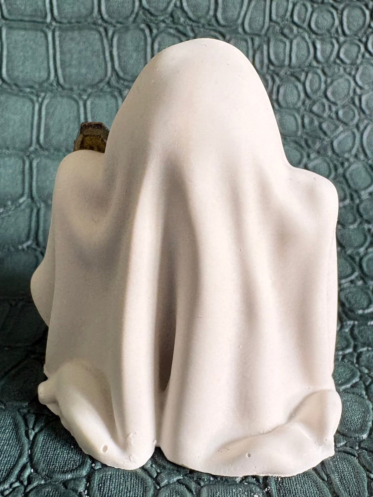 A small white ghost-shaped figurine or candle holder with a draped sheet design, photographed against a dark textured crocodile-embossed background.