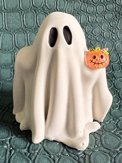 A small white ghost figurine with black oval eyes stands on a textured dark teal surface, holding a tiny orange jack-o'-lantern accessory.