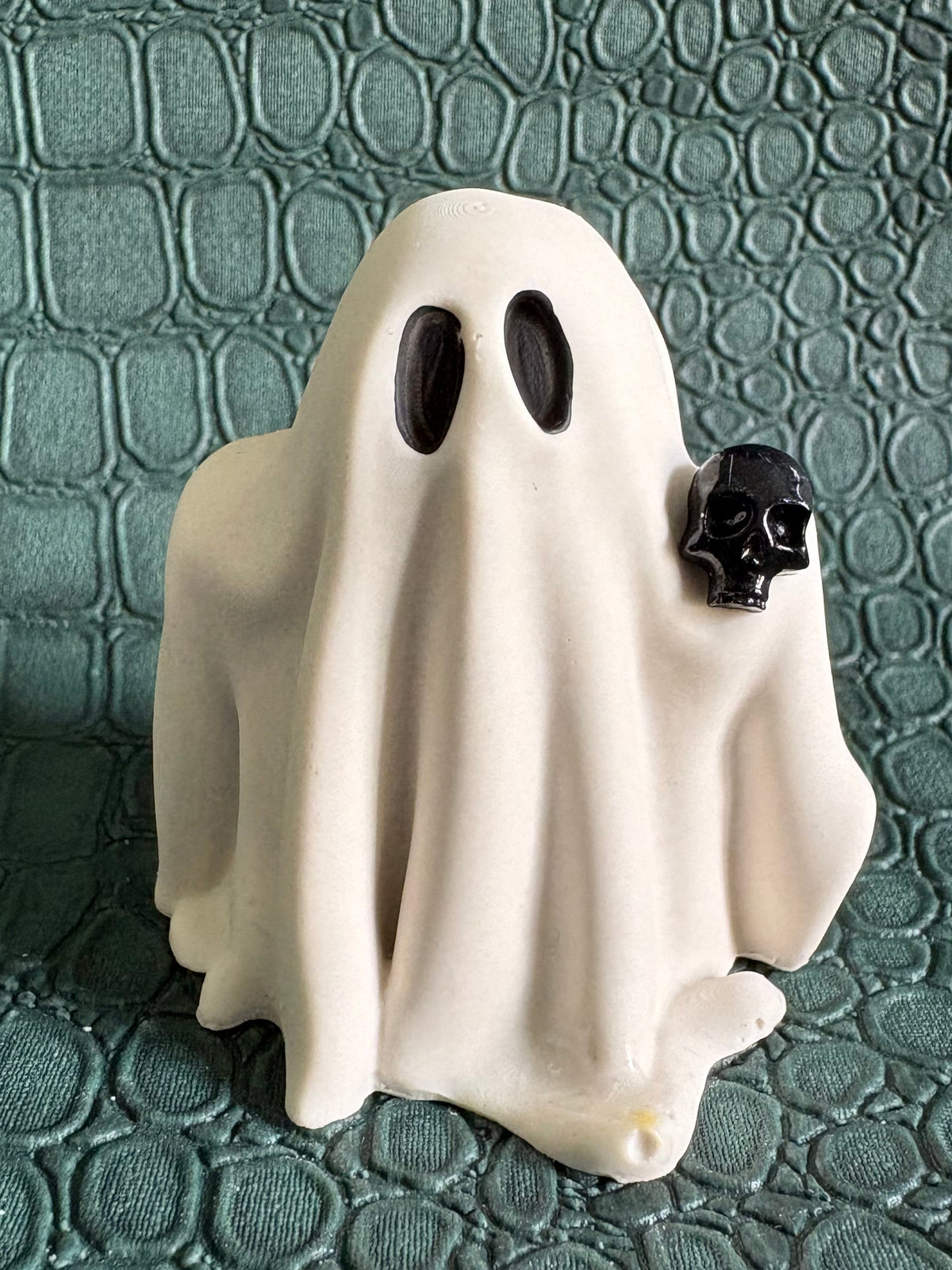 A small white ghost figurine with black eyes and a small black skull accent on the right side, standing on a textured dark green surface.