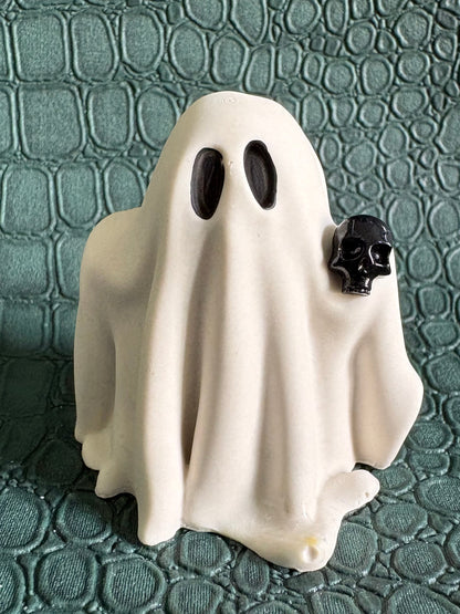A small white ghost figurine with black eyes and a small black skull accent on the right side, standing on a textured dark green surface.