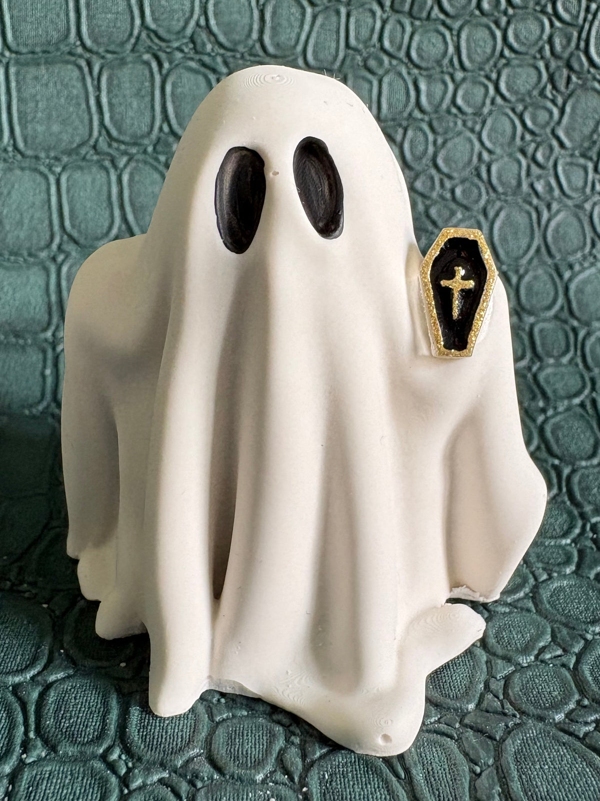 A white ghost-shaped figurine with black oval eyes and a small black coffin-shaped pendant with a gold cross on the right side, set against a textured dark green background.