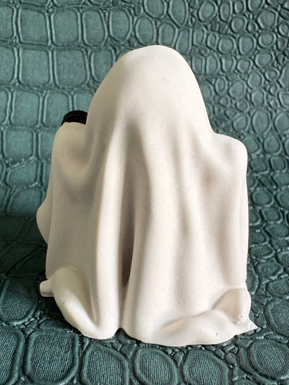 A small white ghost-shaped figurine or candle holder with a draped fabric-like surface, photographed against a textured teal-green background.