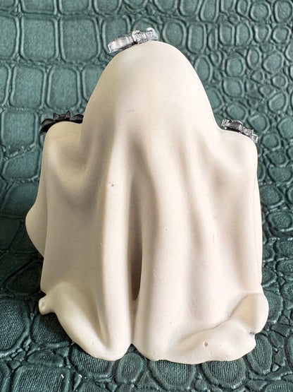 A small cream-colored ghost figurine draped in fabric, sitting on a dark green textured crocodile-patterned surface.