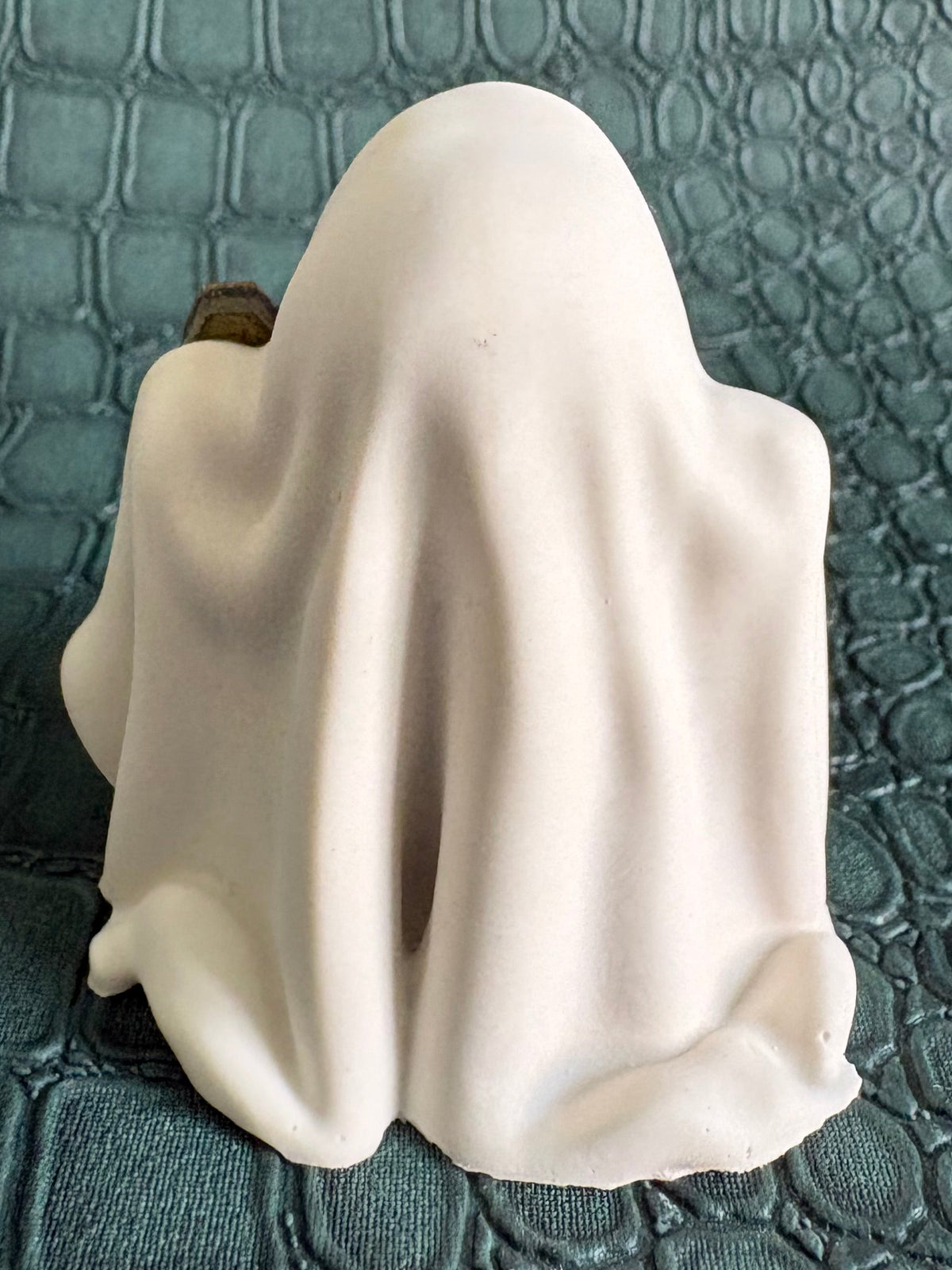 A small white ghost-shaped figurine or candle holder with a draped fabric-like surface, photographed on a dark textured crocodile-embossed background.