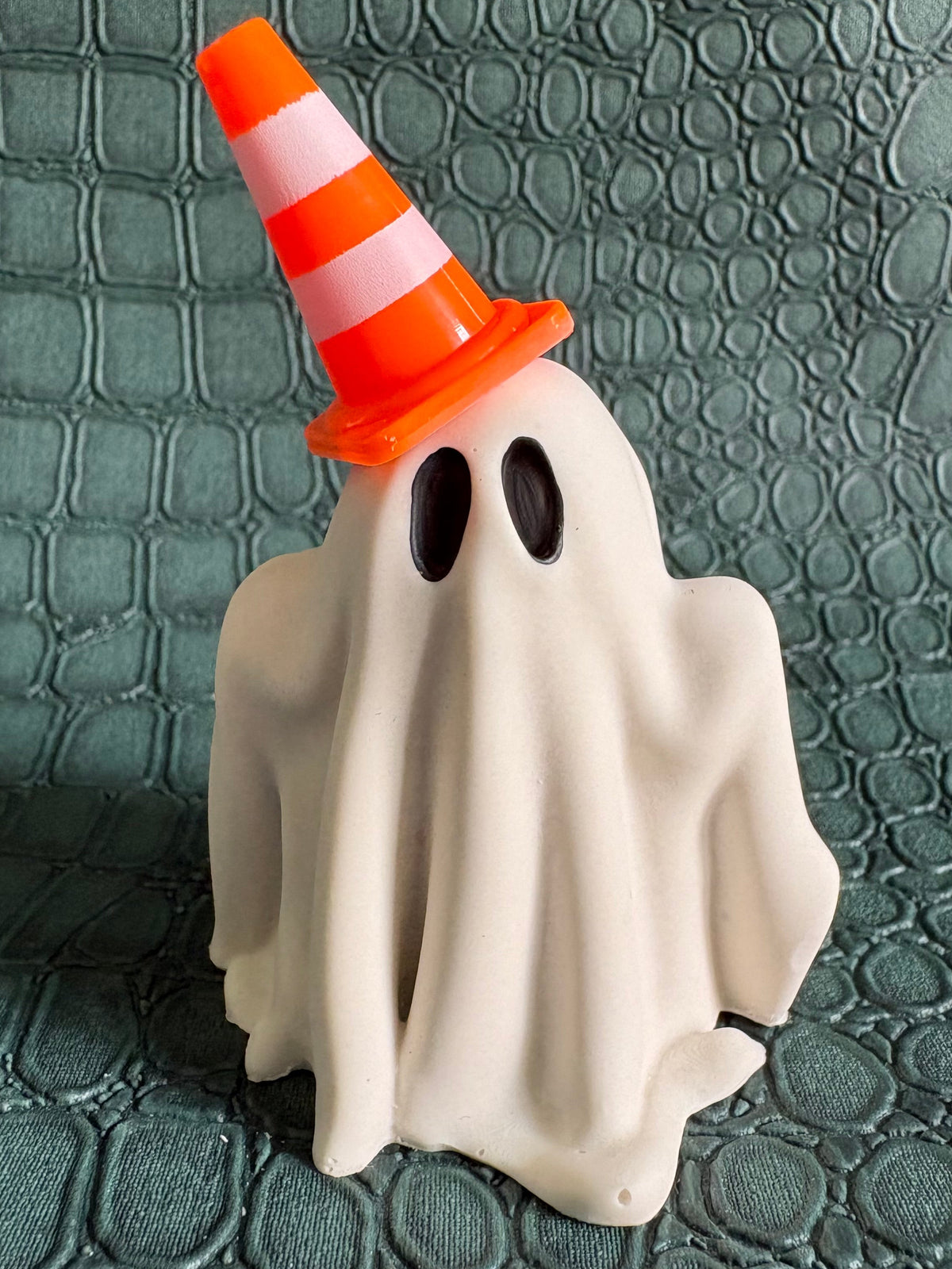 A Halloween ghost decoration shaped like a sheet of white paper with black oval eyes, wearing an orange and white striped traffic cone hat, set against a dark green textured background.