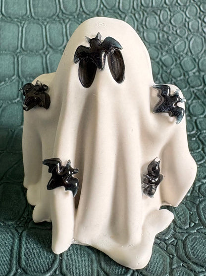 A white ghost-shaped decorative figurine with black bat details, set against a textured dark green crocodile-patterned background.