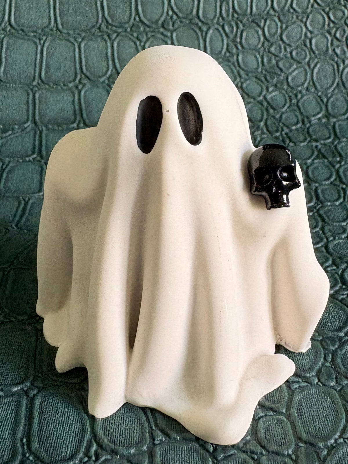 A small white ghost figurine with black oval eyes and a tiny black skull accent on the right side, standing on a dark green textured crocodile-patterned surface.