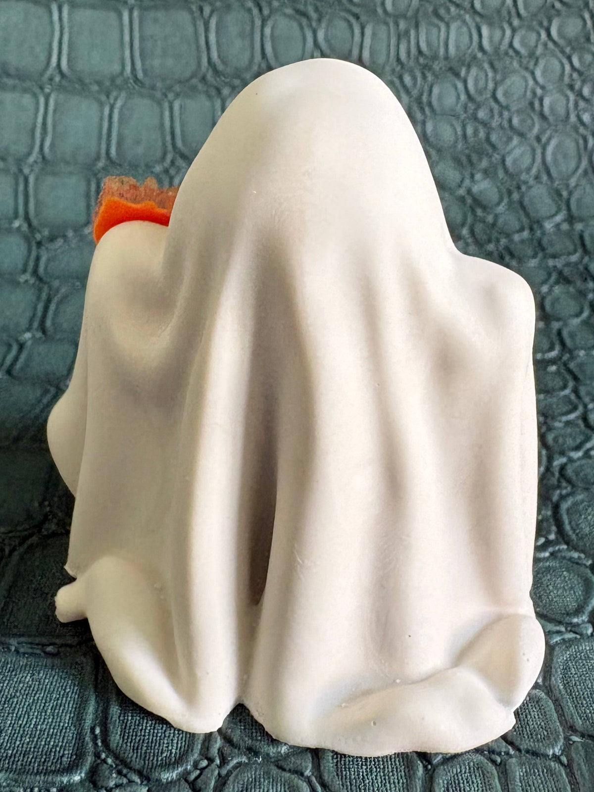 A white ghost-shaped ring or accessory with a small orange accent peeking out from behind it, photographed on a dark textured crocodile-embossed surface.