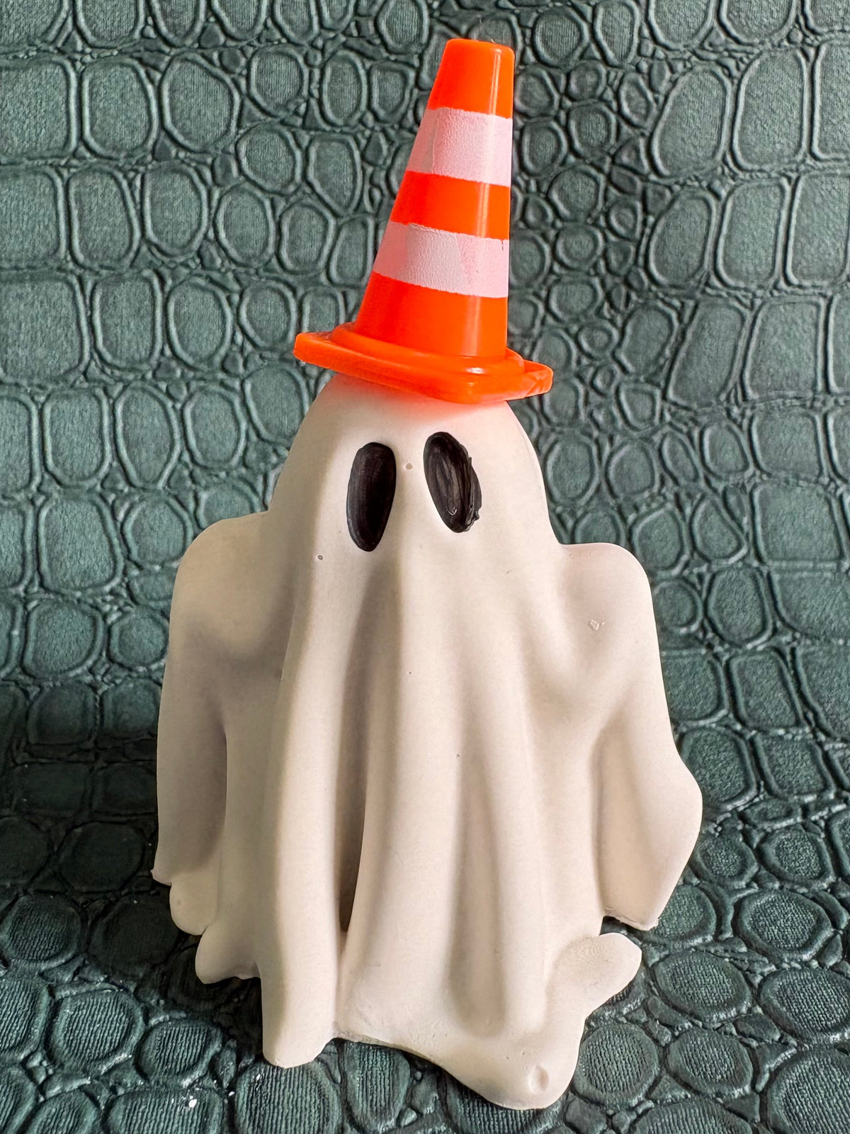 A small Halloween ghost decoration with a white sheet-like body, black oval eyes, and an orange traffic cone hat with white stripes, standing on a textured dark green surface.