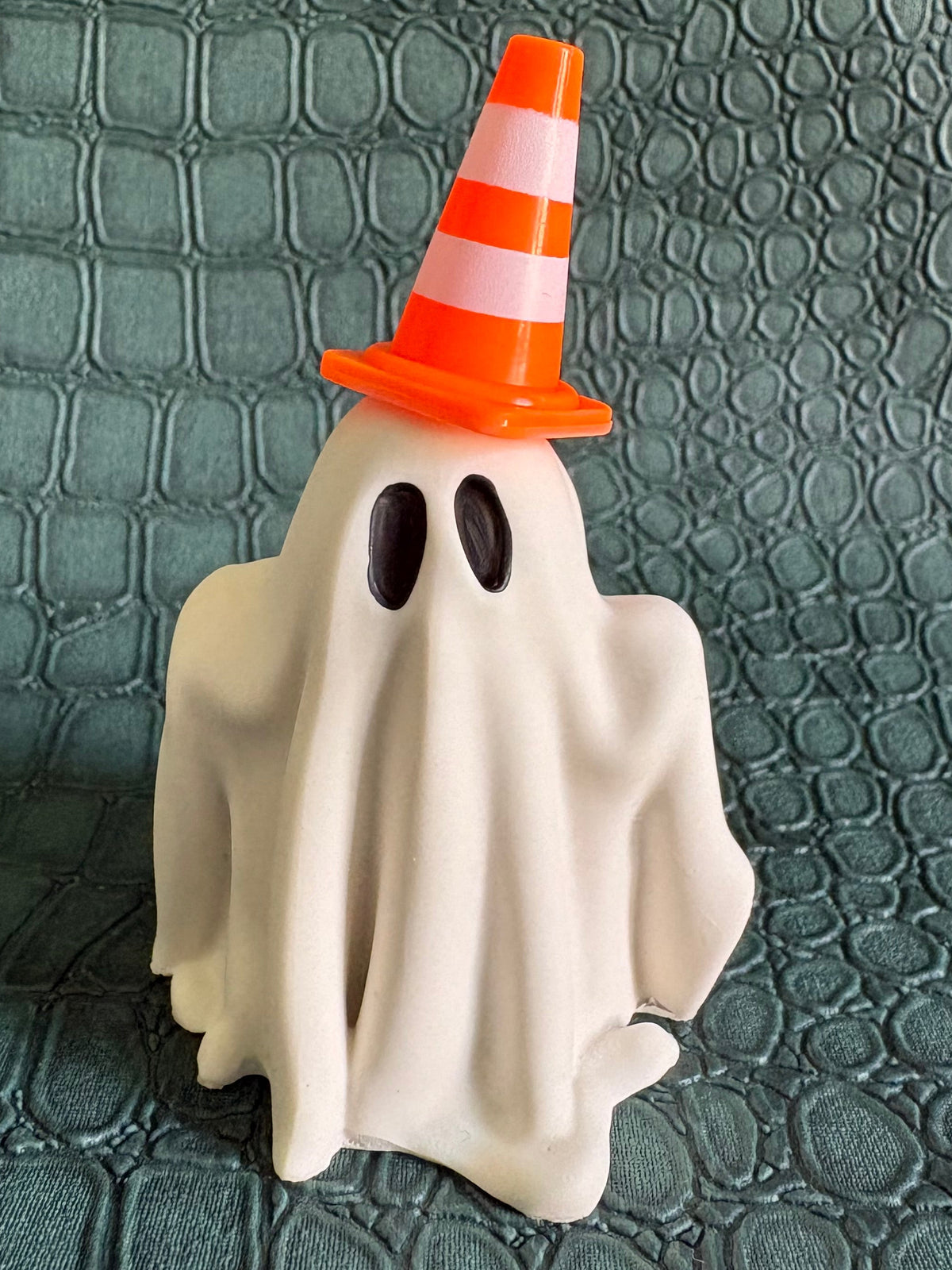 A small Halloween decoration shaped like a ghost draped in white fabric, wearing an orange and white striped traffic cone hat, standing on a textured dark green surface.