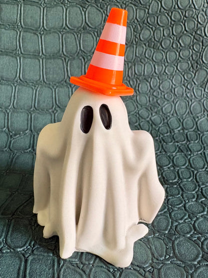 A small Halloween decoration shaped like a ghost draped in white fabric, wearing an orange and white striped traffic cone hat, standing on a textured dark green surface.