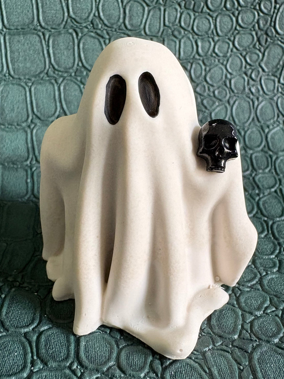A small white ghost-shaped figurine with two black oval eyes and a small black skull detail on the right side, standing on a textured dark green surface.