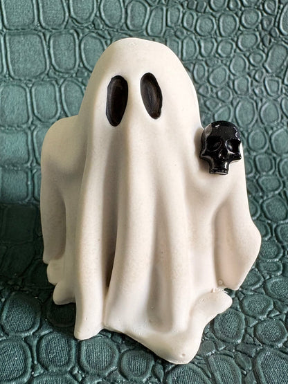 A small white ghost-shaped figurine with two black oval eyes and a small black skull detail on the right side, standing on a textured dark green surface.