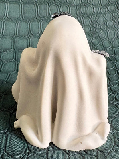 A small ghost-shaped figurine or candle holder in an off-white fabric-like drape, photographed from the back on a textured dark green crocodile-embossed surface.
