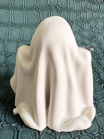 A small white ghost-shaped figurine or candle holder draped in fabric, photographed against a dark textured crocodile-pattern background.