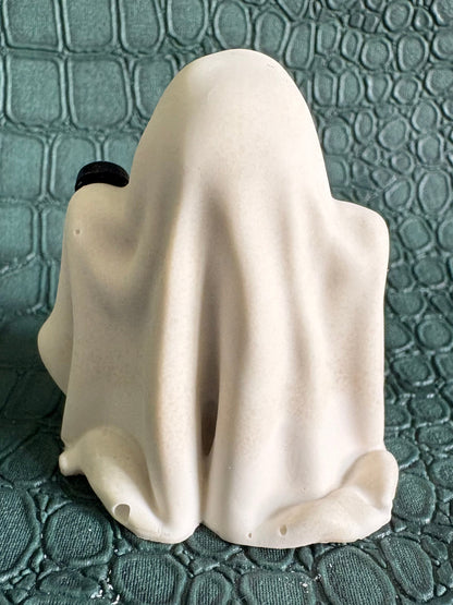 A small ghost-shaped figurine draped in a white sheet, photographed against a textured dark teal crocodile-embossed surface.