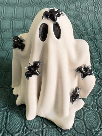 A white ghost-shaped figurine with black bat details and black eye cutouts, displayed on a textured dark green surface.