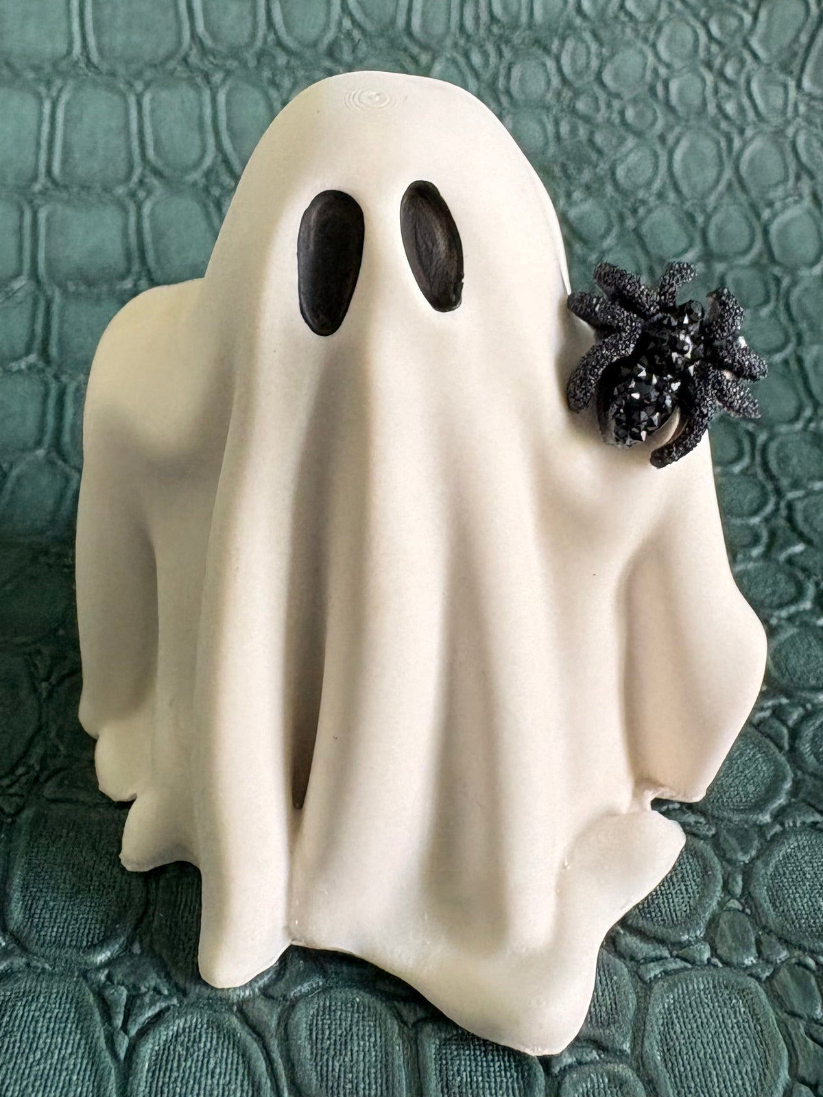 A small white ghost figurine with black eyes and a black spider decoration on its back, standing on a textured dark green surface.
