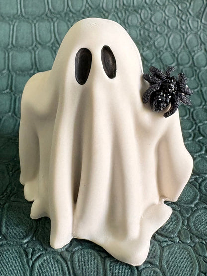 A small white ghost figurine with black eyes and a black spider decoration on its back, standing on a textured dark green surface.