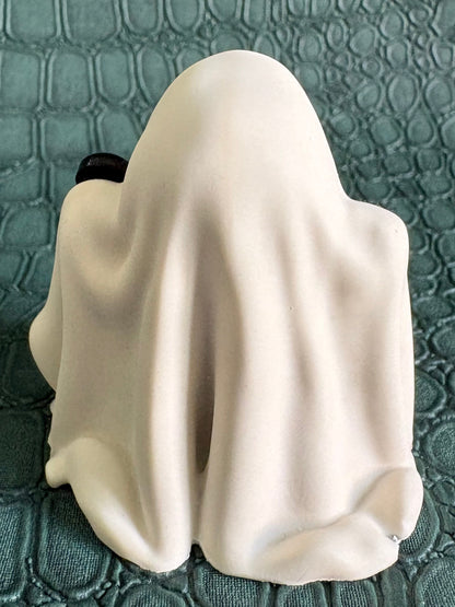 A small white ghost-shaped figurine or candle holder with a draped sheet design, photographed against a textured dark green crocodile-embossed surface.