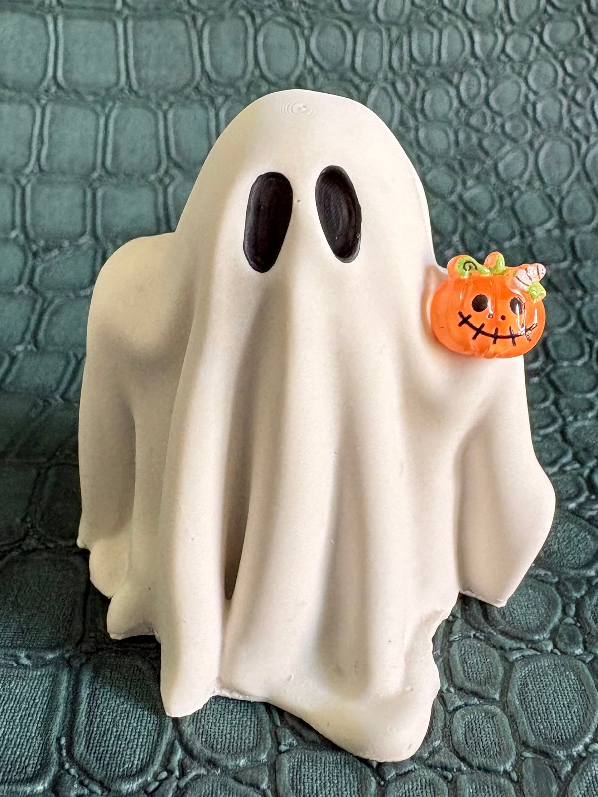 A small white ghost figurine with black eyes stands on a textured dark green surface, holding a tiny orange jack-o'-lantern accent.