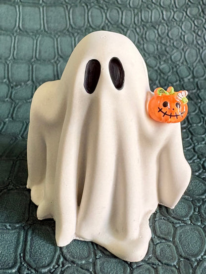 A small white ghost figurine with black eyes stands on a textured dark green surface, holding a tiny orange jack-o'-lantern accent.