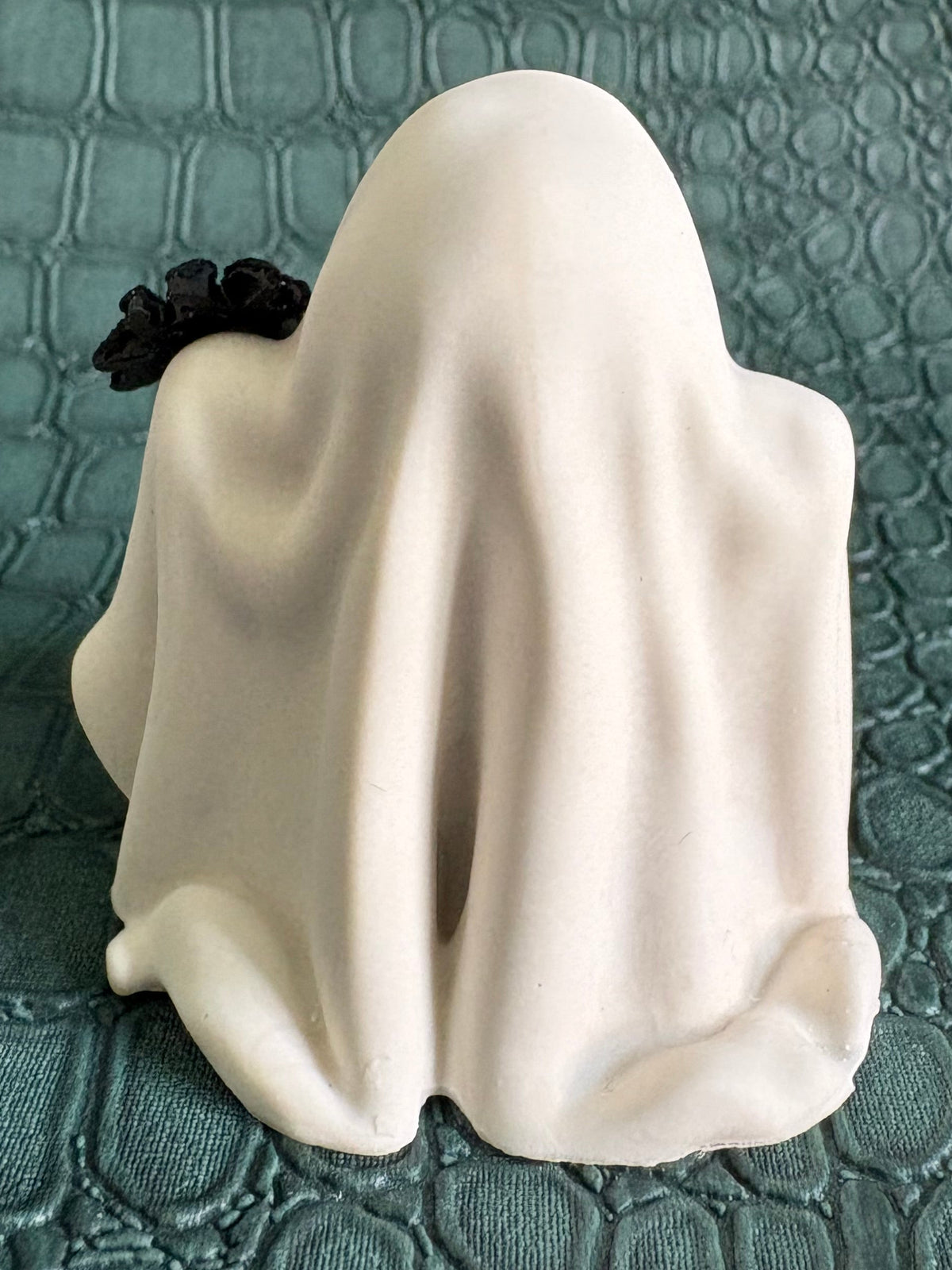 A small white ghost figurine draped in a sheet, with a black scrunchie on its head, standing on a textured teal surface.