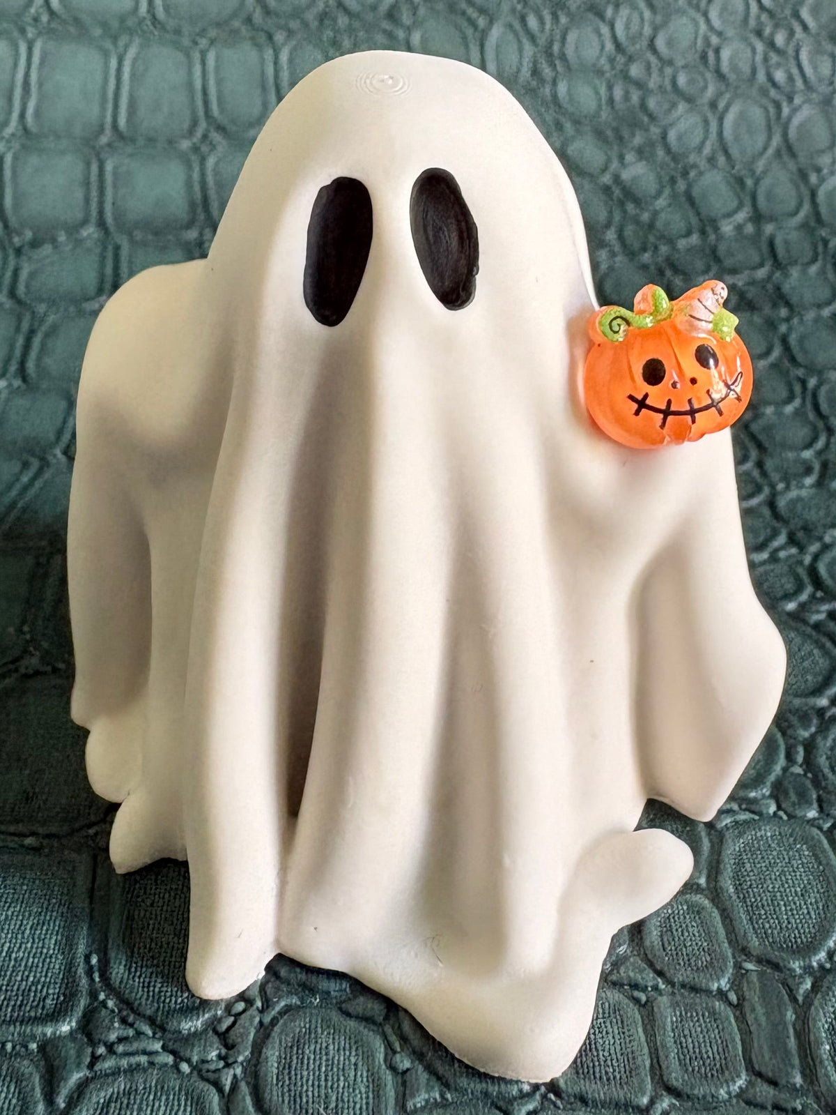 A white ghost-shaped figurine with black oval eyes holding a small orange jack-o'-lantern, set against a dark textured crocodile-patterned background.