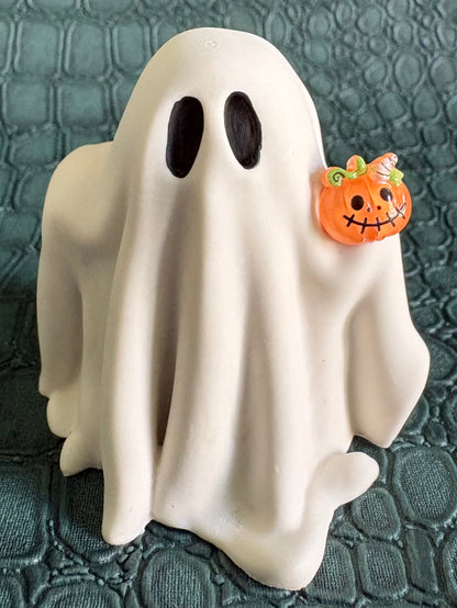 A white ghost-shaped figurine with black oval eyes holding a small orange jack-o'-lantern, set against a dark textured crocodile-patterned background.