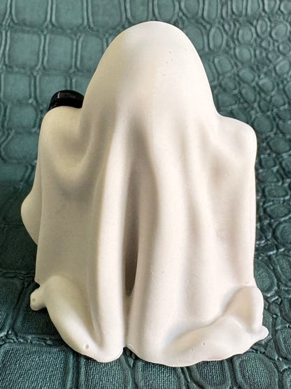 A small white ghost-shaped figurine or candle holder with a draped fabric-like surface, photographed from the back on a textured teal surface.