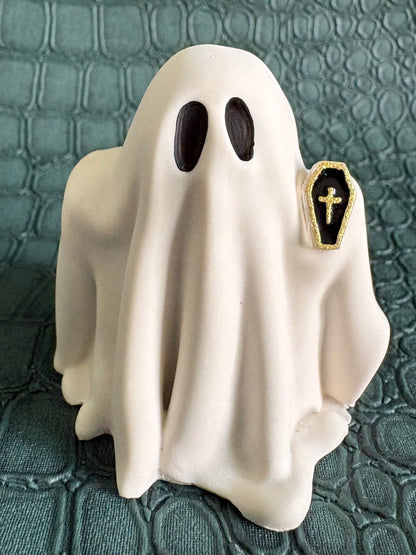 A small white ghost figurine with black oval eyes and a small black coffin-shaped cross charm on the side, photographed against a dark textured crocodile-embossed background.