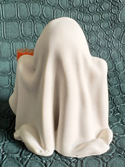 A white ghost-shaped figurine or candle with a draped sheet design, positioned against a textured teal-green background.