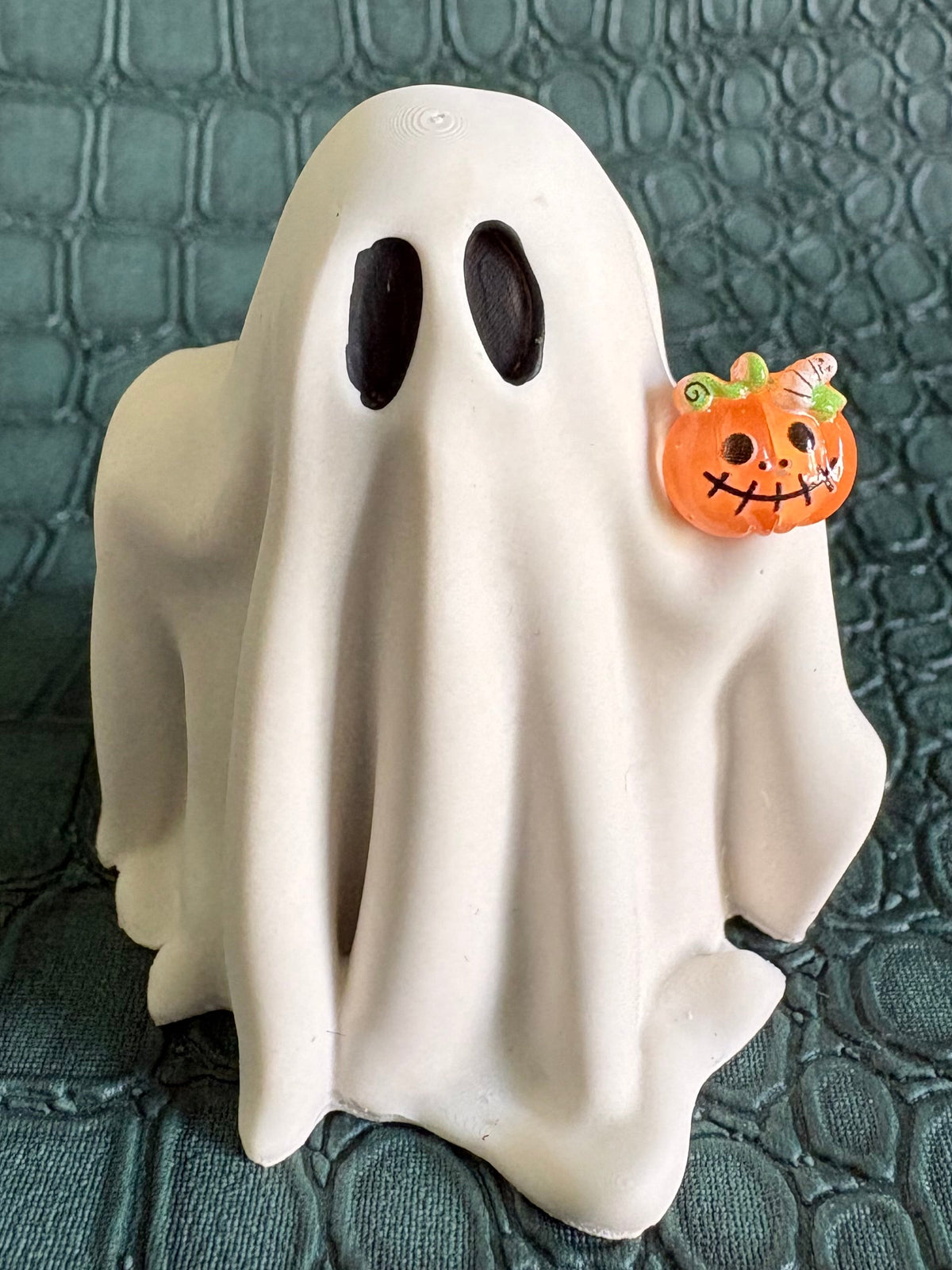A white ghost-shaped Halloween decoration with black oval eyes and a small orange jack-o'-lantern accent on the right side, standing on a dark textured surface.