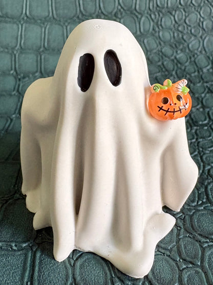 A white ghost-shaped Halloween decoration with black oval eyes and a small orange jack-o'-lantern accent on the right side, standing on a dark textured surface.