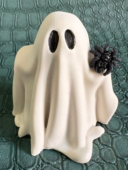 A small white ghost figurine with black eyes and a black spider decoration on its right side, standing on a textured dark green surface.