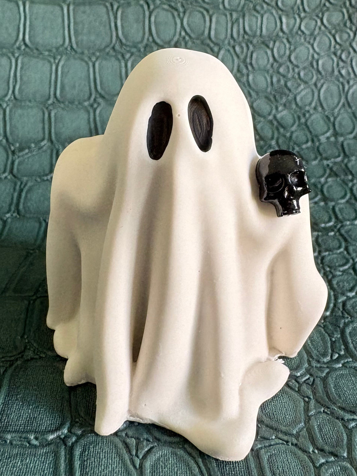 A small white ghost figurine with black oval eyes and a small black skull accent on the right side, standing on a textured teal surface.