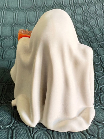 A small white ghost-shaped figurine or candle holder with a draped fabric-like surface, photographed on a dark textured green surface.