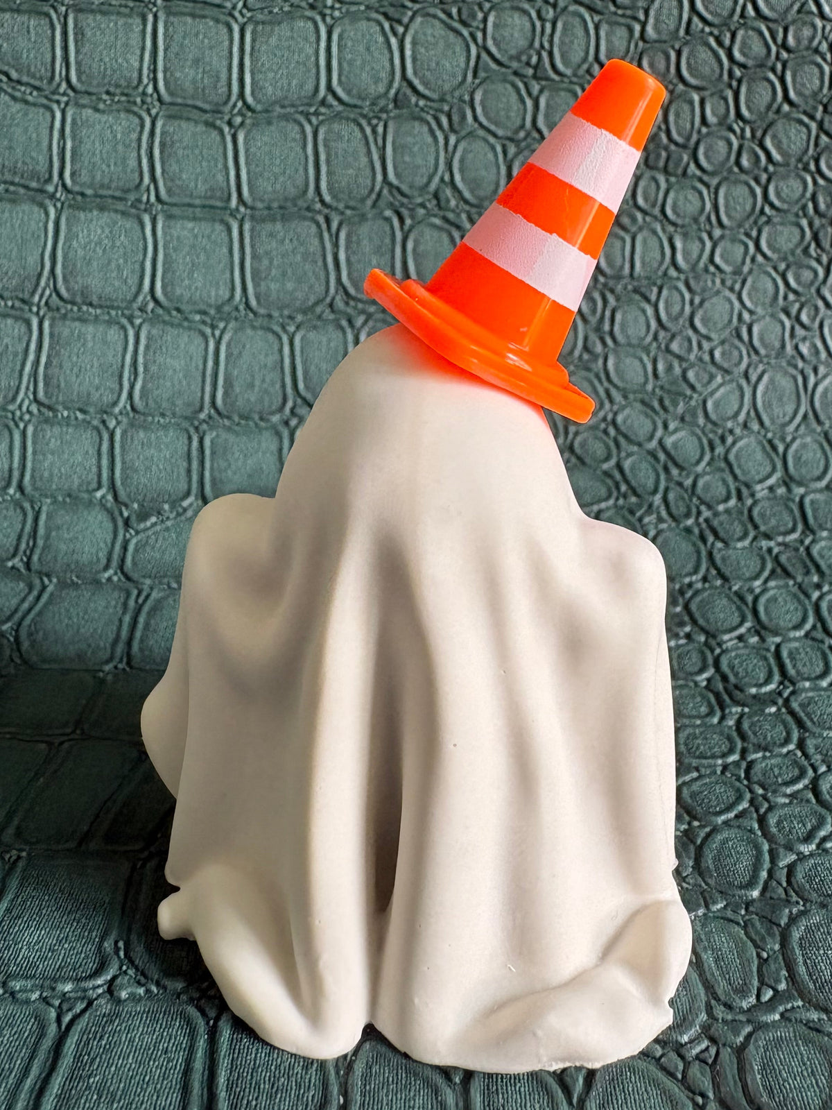 A Halloween decoration shaped like a ghost draped in a white sheet, wearing an orange and white striped traffic cone hat, set against a dark green textured crocodile-patterned background.