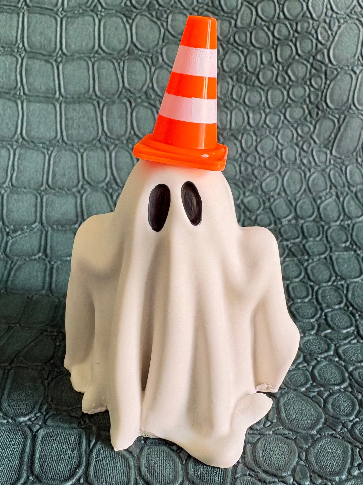 A small Halloween ghost figurine draped in a white sheet, wearing an orange and white striped traffic cone hat, standing on a textured dark green surface.