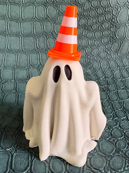 A small Halloween ghost figurine draped in a white sheet, wearing an orange and white striped traffic cone hat, standing on a textured dark green surface.