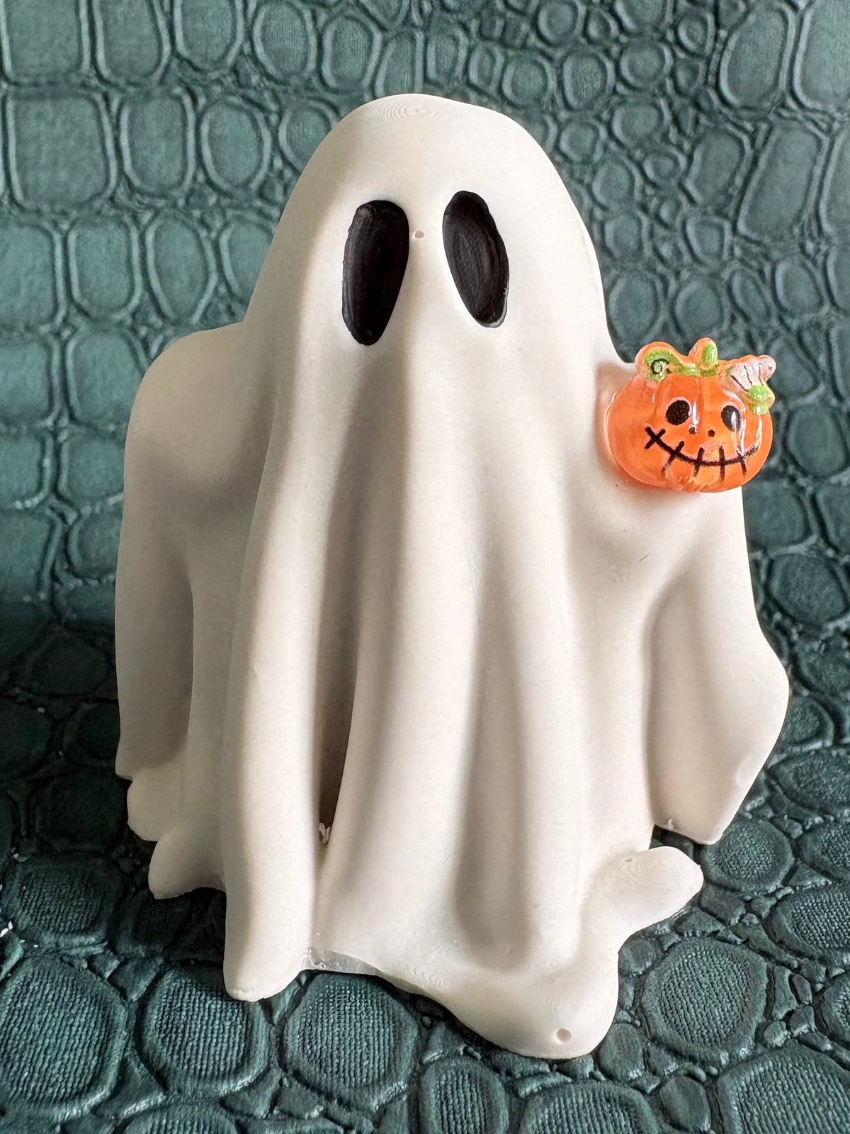 A white ghost-shaped figurine with black eyes stands on a textured dark green surface, holding a small orange jack-o'-lantern charm.