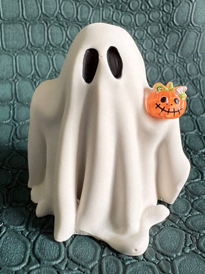 A white ghost-shaped figurine with black eyes stands on a textured dark green surface, holding a small orange jack-o'-lantern charm.